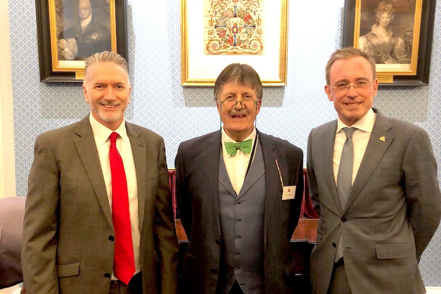 Three men in suits stand in front of paintings.