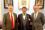 Three men in suits stand in front of paintings.