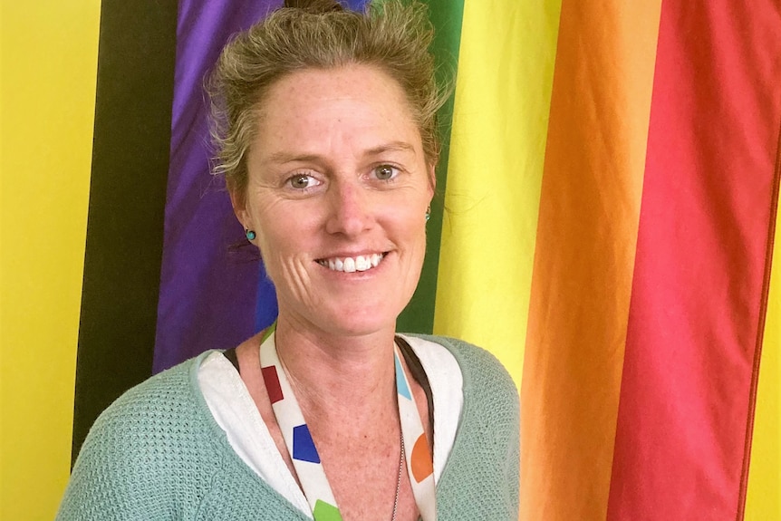 A woman with tied back brown hair standing in front of rainbow flag smiling
