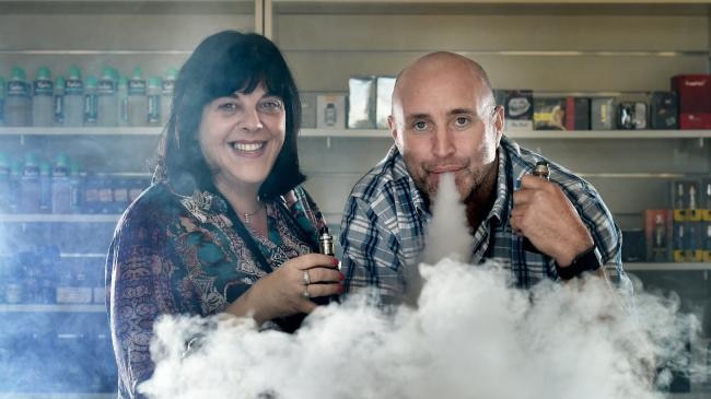 A woman and a man pose for the camera in front of a counter of tobacco vaping products. The man exhales a cloud of vapor