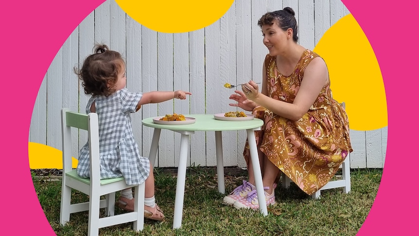 A toddler sits at an outdoor kids table while her mum tries to feed her dinner, the challenges of getting toddlers to eat.