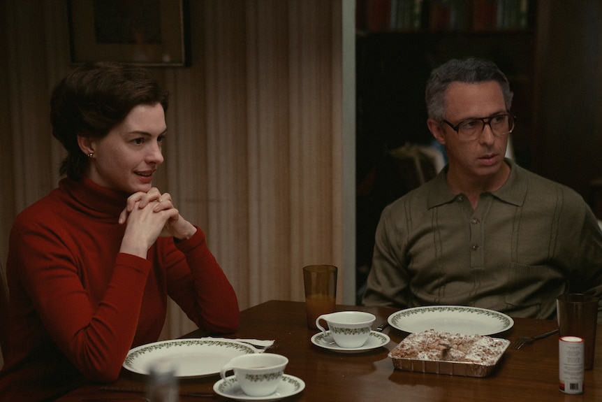 餐桌旁坐着一位身穿红色长袖衬衫的白褐发女郎，旁边是一位戴着眼镜和卡其色衬衫的灰白男人。