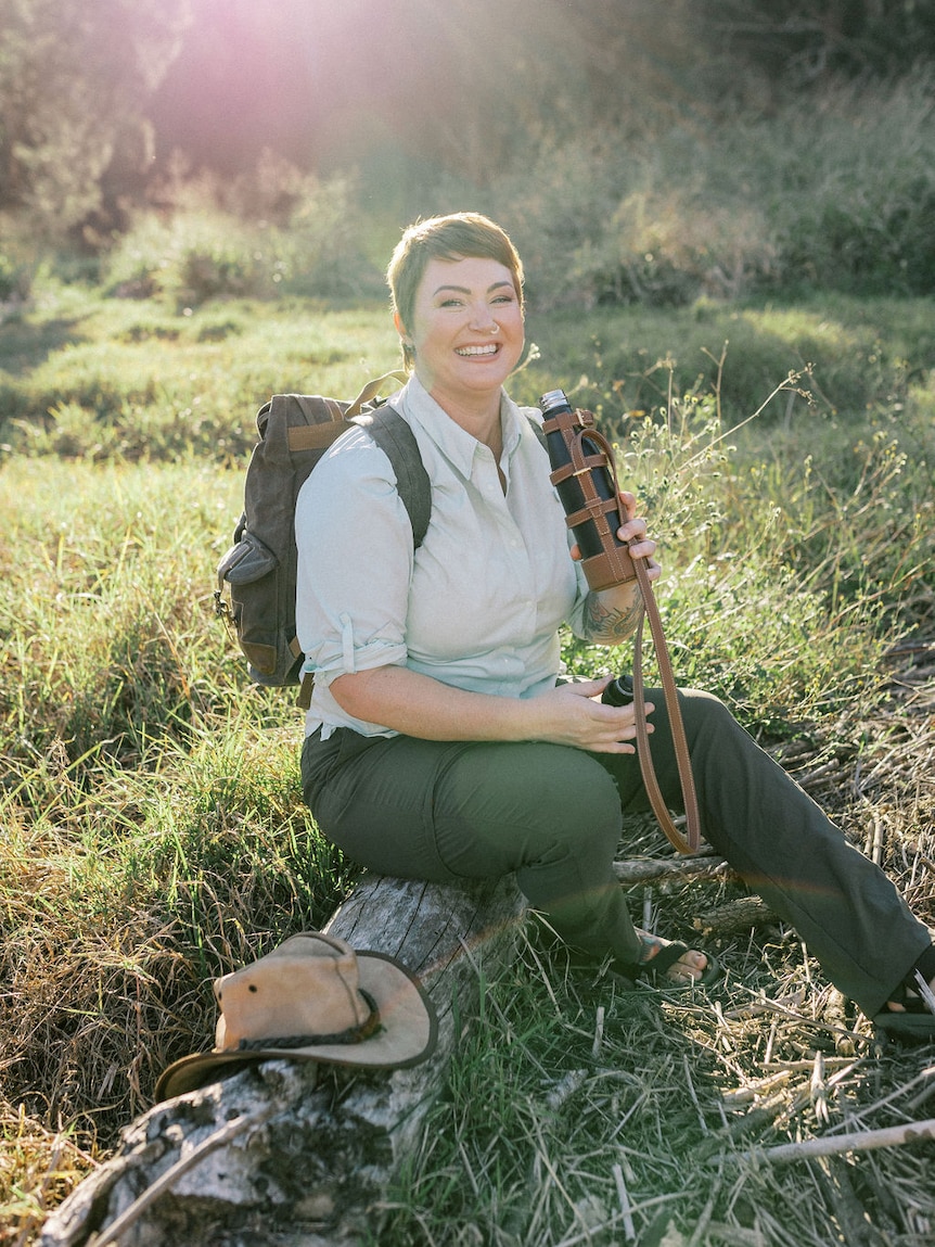 A woman in shirt and pants, sitting on a log, drinking from a water bottle, smiles at the camera 