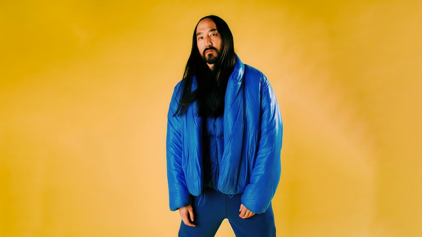 Steve Aoki wears a blue puffer jacket with blue shirt & pants. He has black long hair & facial hair. Yellow background.