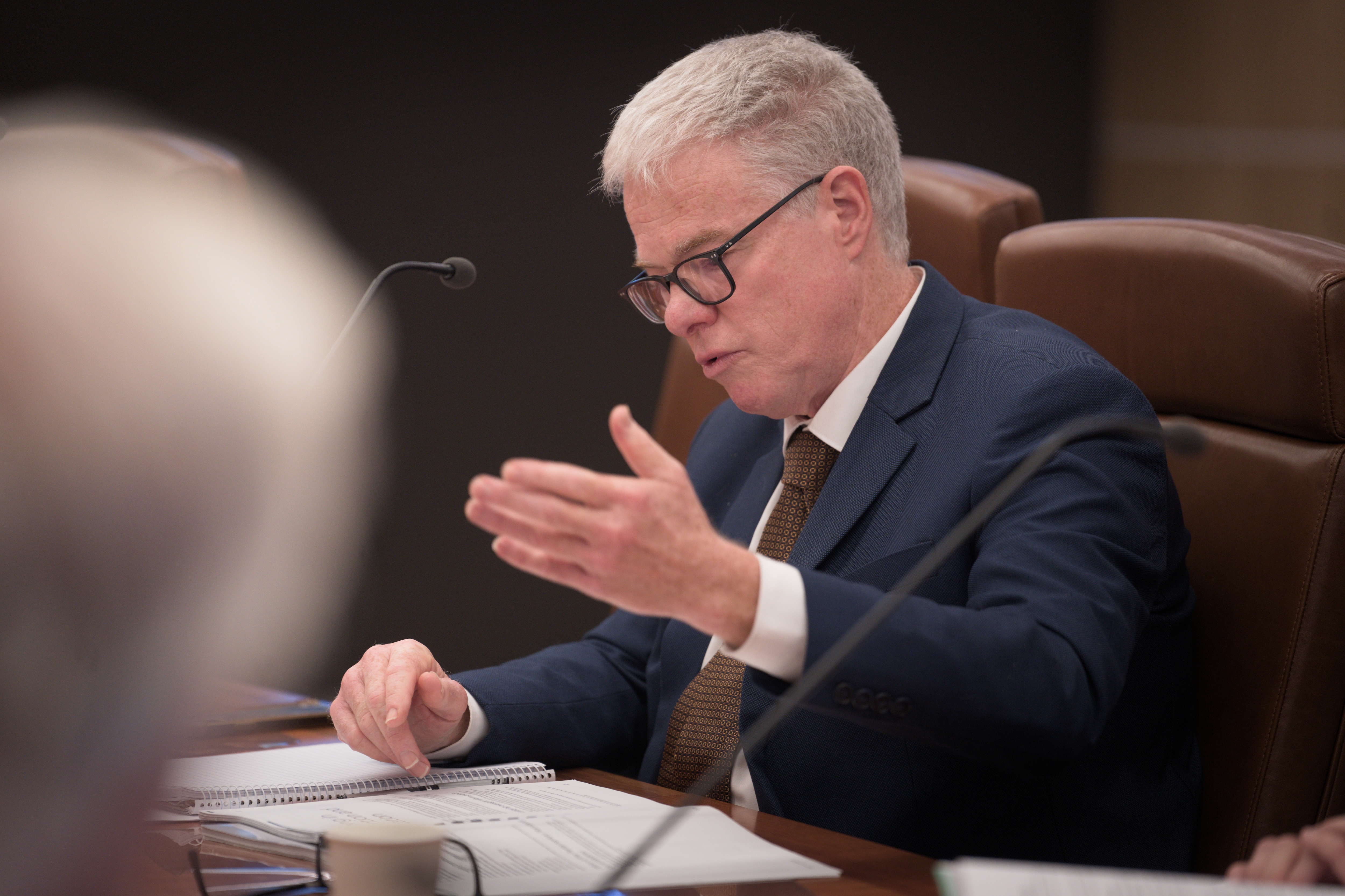 A middle aged man in a dark suit and glasses reads off paper