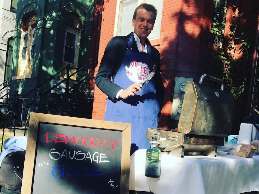 Rowland wearing apron holding tongs and standing behind a BBQ with a sign saying Democracy Sausage Sizzle.