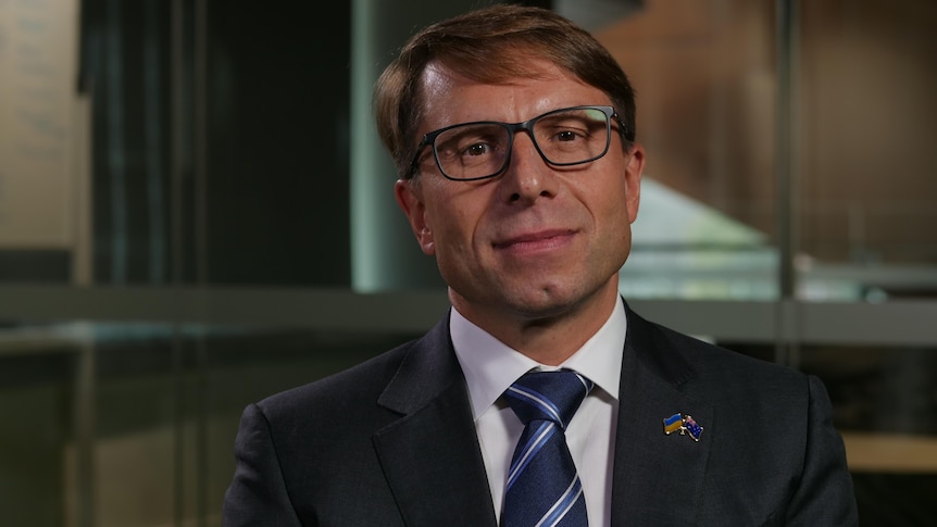 A man with short, dark hair and spectacles, wearing a dark suit.