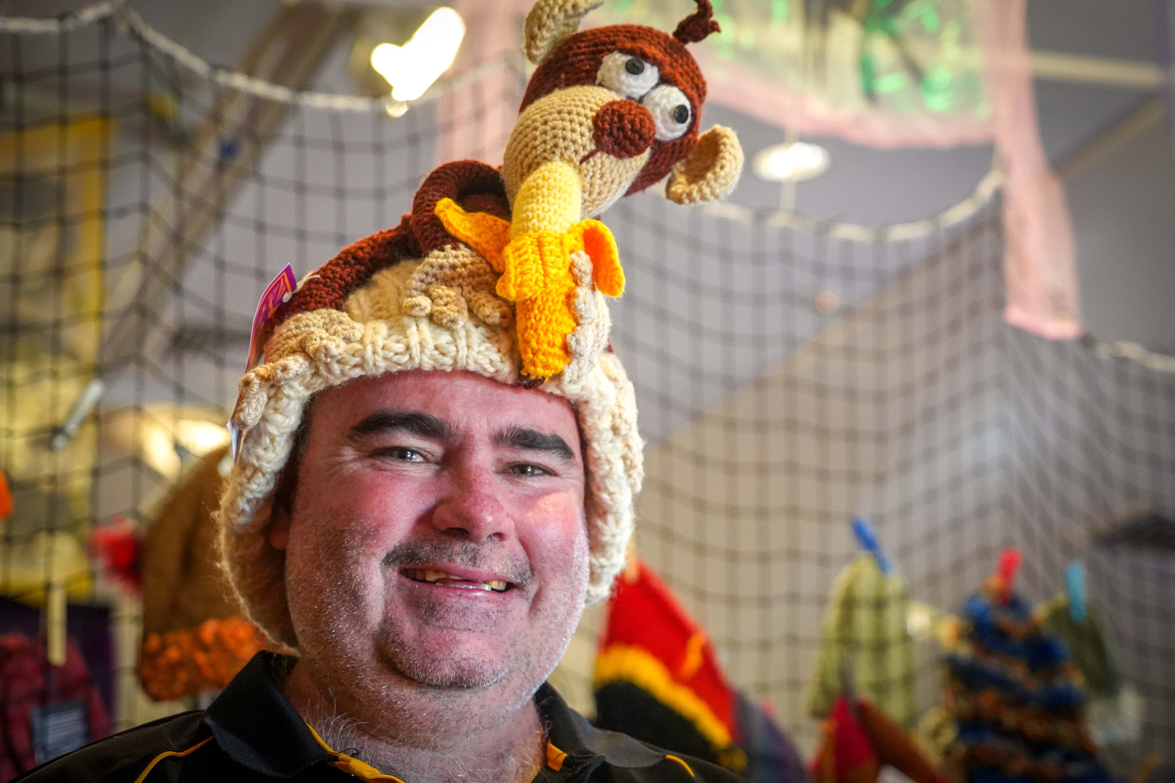 A man smiling at the camera, on his head is a handmade beanie featuring a knitted monkey holding a banana.