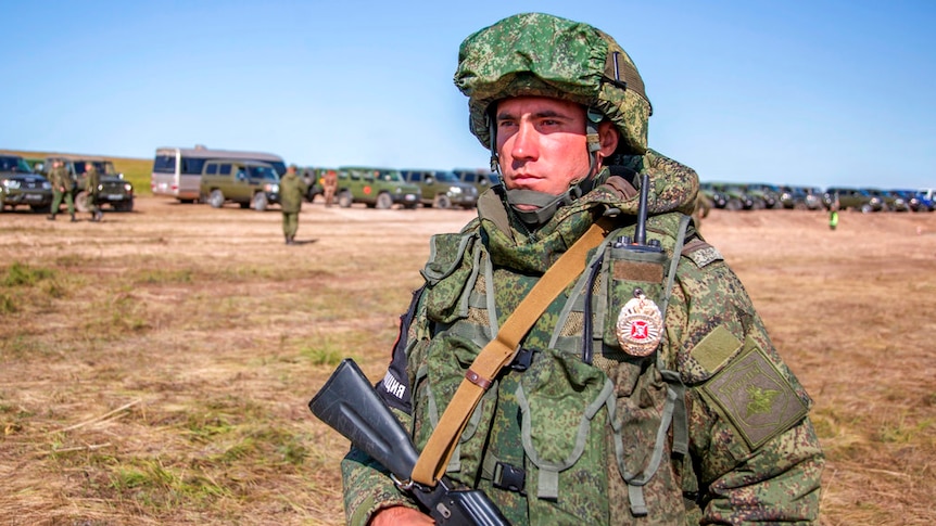 A soldier with a gun wearing camouflage gear and helmet, with other soldiers and army vehicles lined up behind him