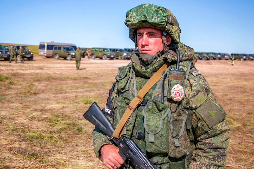 A soldier with a gun wearing camouflage gear and helmet, with other soldiers and army vehicles lined up behind him