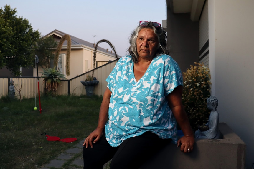 A woman sits on a planter outside a house. She looks off, with a neutral expression. The yard behind her has a totem tennis set.