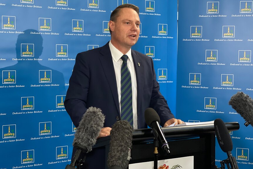 Brisbane Lord Mayor Adrian Schrinner wears a suit and blue tie while speaking in front of microphones