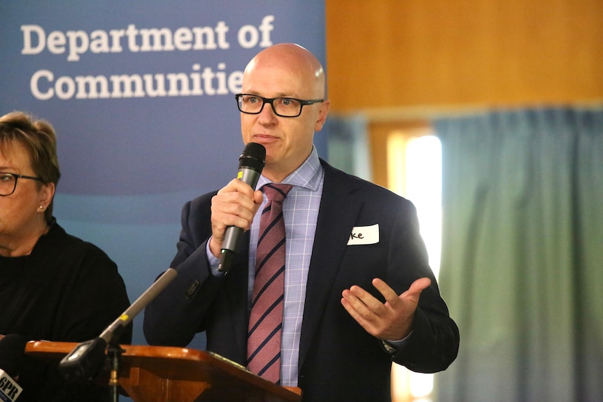 A bald man wearing a suit and glasses holds a microphone while speaking at a lectern