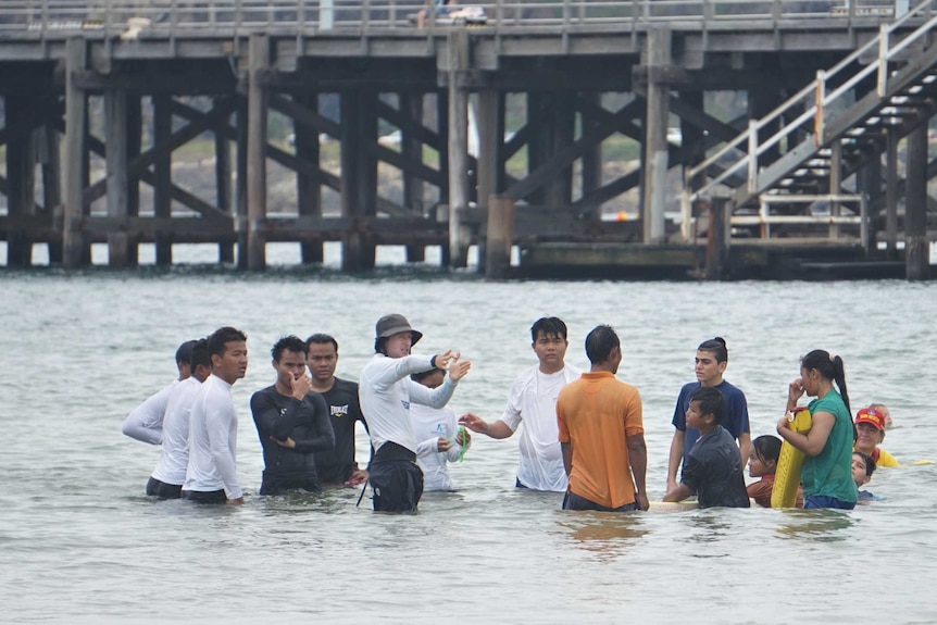 15 people in thigh deep sea water near large timber pier listen to swimming instructions