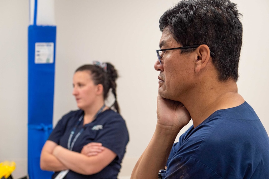 Two healthcare workers in blue scrubs, a man in the foreground rubs his neck.
