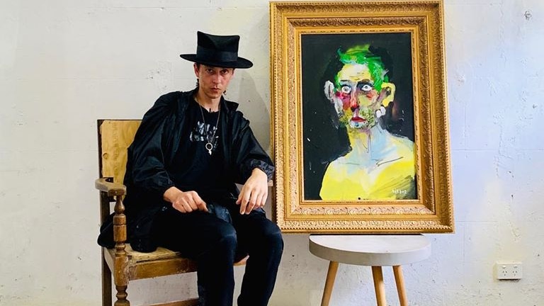 Anthony Lister sits wearing hat next to a painting of a man