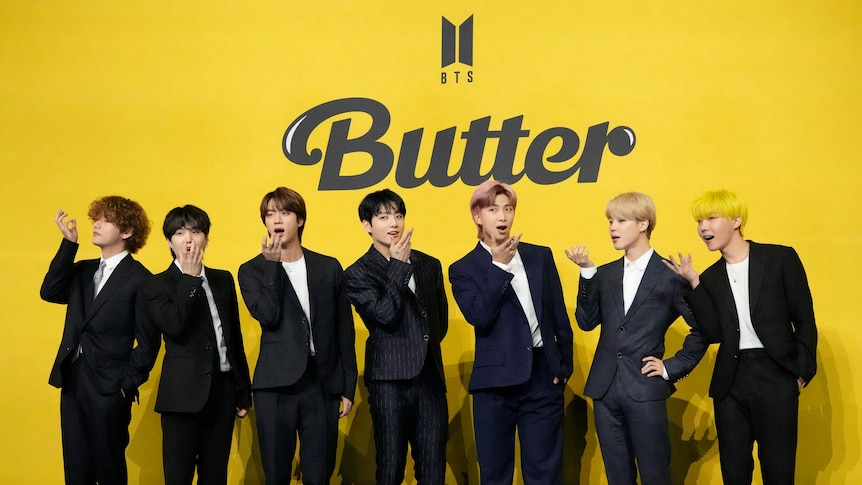Seven trendy-looking young Korean men in suits pose in front of a yellow background.