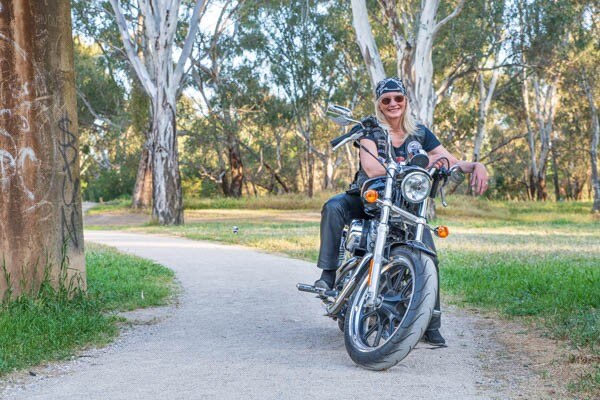 A blonde woman in a black bandana poses on a silver and black motorcycle in a park.