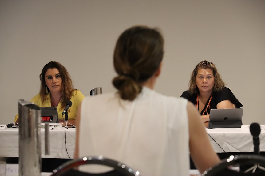 Two women sitting at a long table with microphones look ahead seriously. 