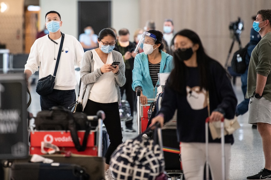 Passengers wearing masks and wheeling their luggage at an airport arrivals terminal.