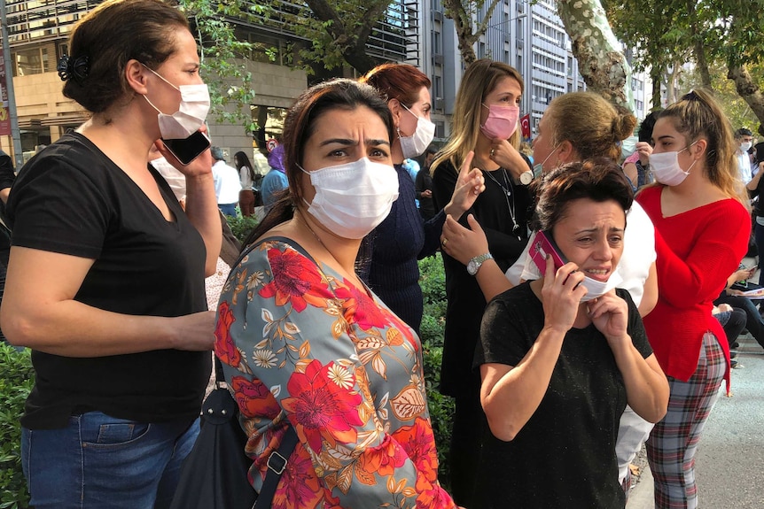 A group of women wearing masks and with concerned looks on their faces stand on a street, some talking on phones