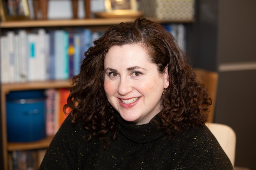 A smiling woman with dark curly hair and a dark jumper sits in front of shelves of books.