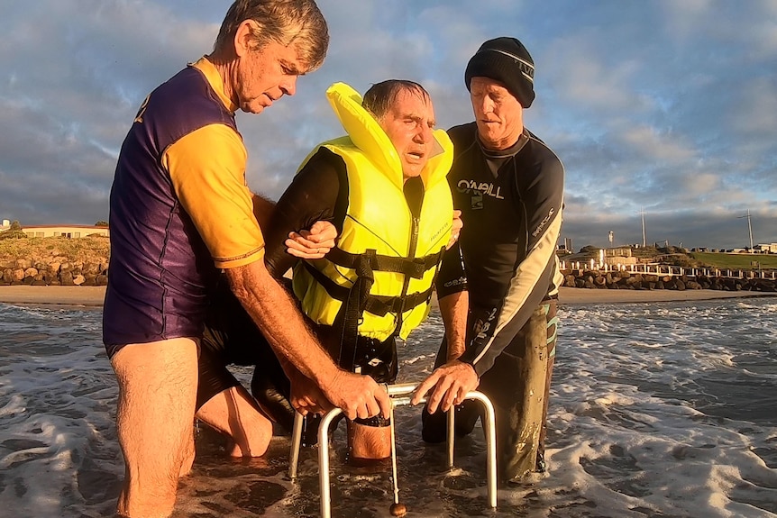 Man in ocean with life jacket with walking frame and two friends helping 