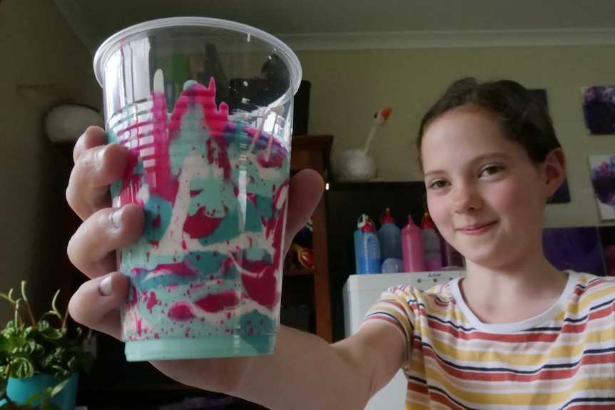 Young girl smiling as she holds up a plastic cup filled with bright pink and aqua coloured paint
