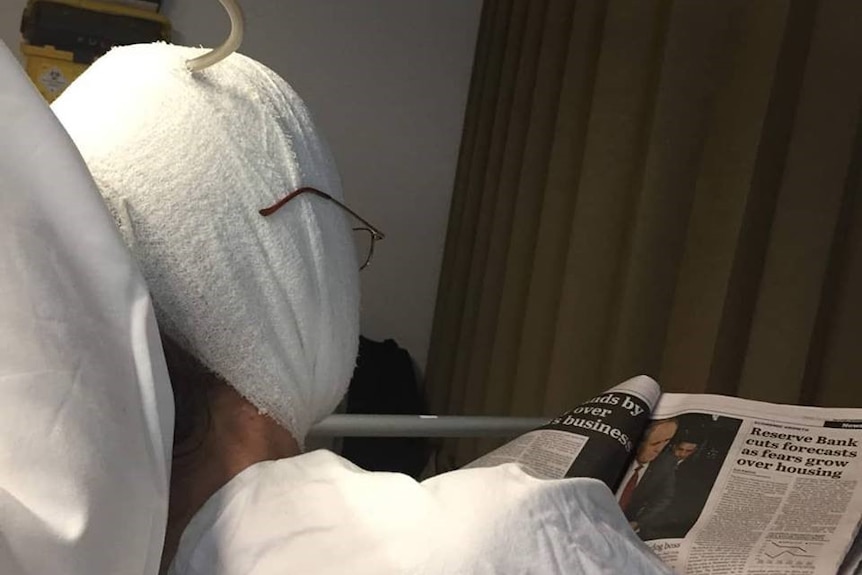 A patient lies in a hospital bed with head wrapped in bandages, reads newspaper