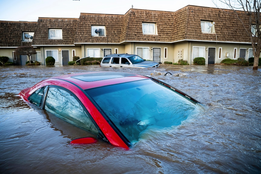 A red car is submerged in floodwater in front of a house. 