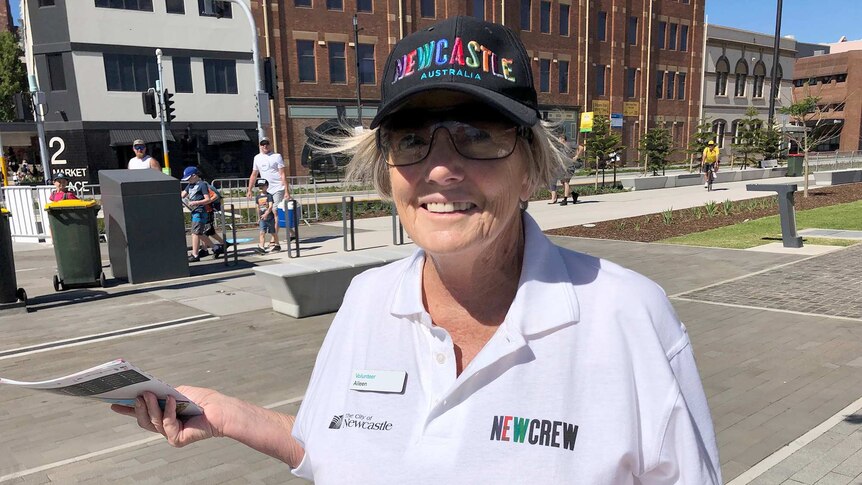 An older woman with a Newcastle hat hands out maps to Supercars visitors.