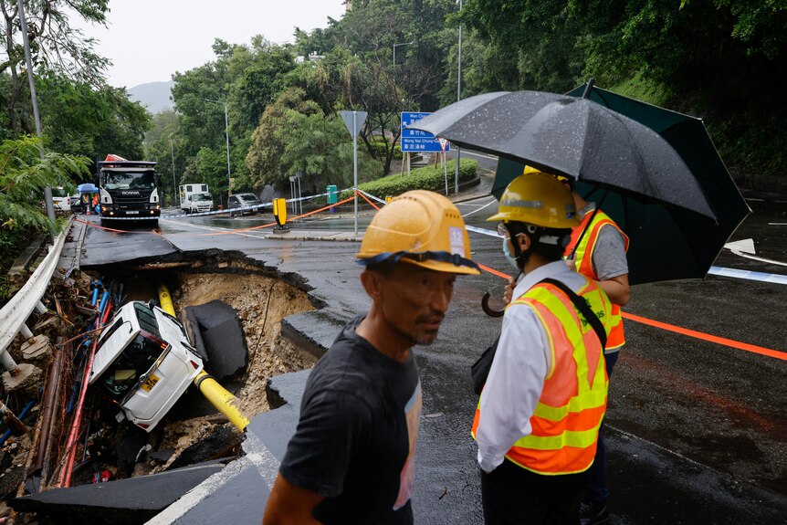 Workers wearing yellow helmets and holding black umbrellas surrounding a large hole in a road with a white car fallen inside