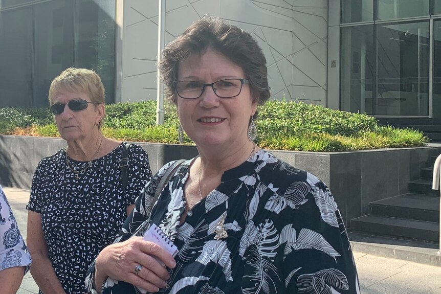 Kathy Phelan wearing a black and white floral shirt, standing outside the WA District Court building with another lady behind.