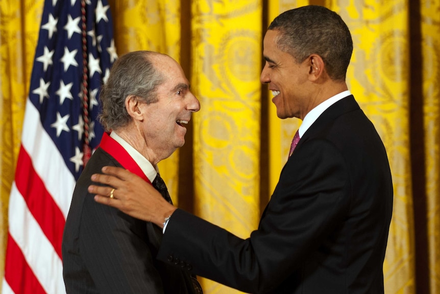 Roth and Obama face each other, smiling warmly, Roth wearing a red ribbon, in front of an ornate yellow curtain.