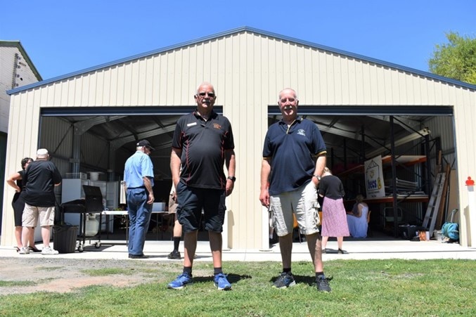 Two men in shorts and dark shirts stand smiling in front of a large, open shed with people milling inside and outside it.