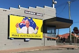 A grey suburban building with a bright yellow sign saying Make Australia Great, with a photo of Clive Palmer and flag.