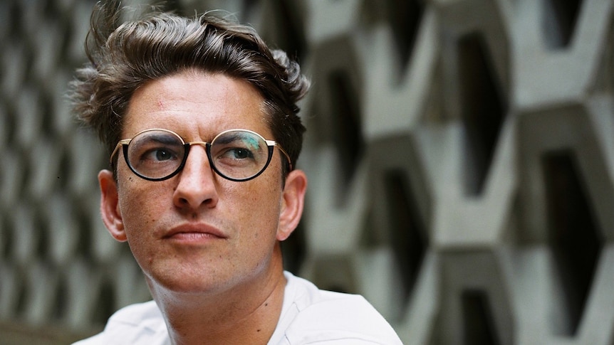 Skream has short brown hair & wears a white tee & black and gold framed glasses. He is looking right, serious facial expression.