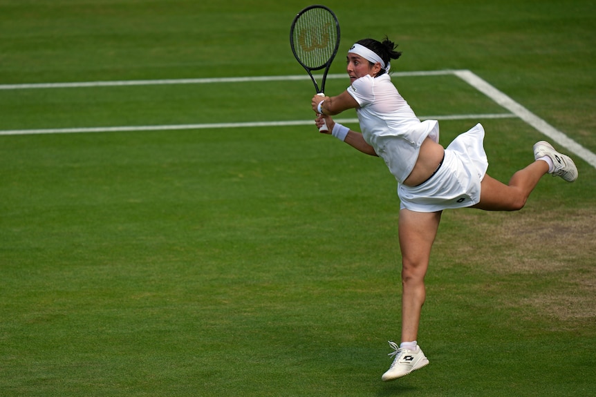 A woman in white hits a high back hand on a grass court.