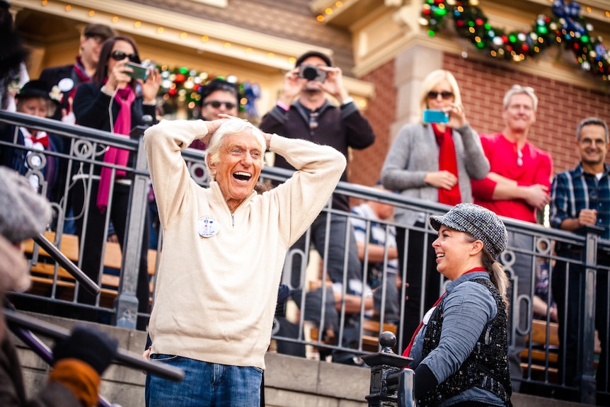 An older man stands with hands on head, looking over his 90th birthday celebrations at Disneyland, his younger wife at his right