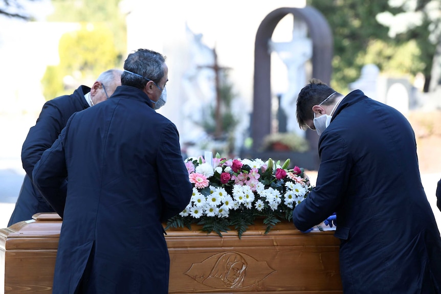 Three men in face masks handling a coffin covered in flowers