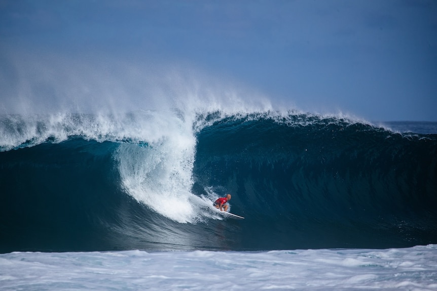 A man in a red singlet grabs the rail of his surfboard as he drops down the face of a huge wave.