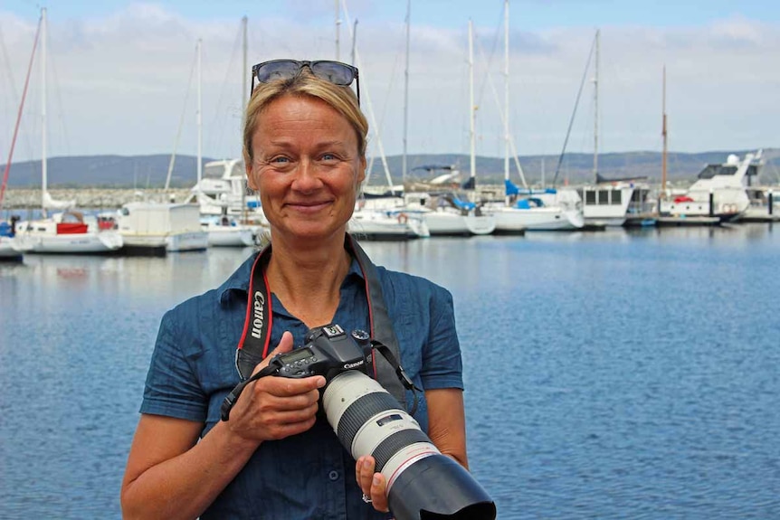 A woman with blonde hair stands in front of a marina holding a large camera.