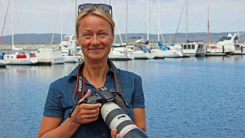 A woman with blonde hair stands in front of a marina holding a large camera.