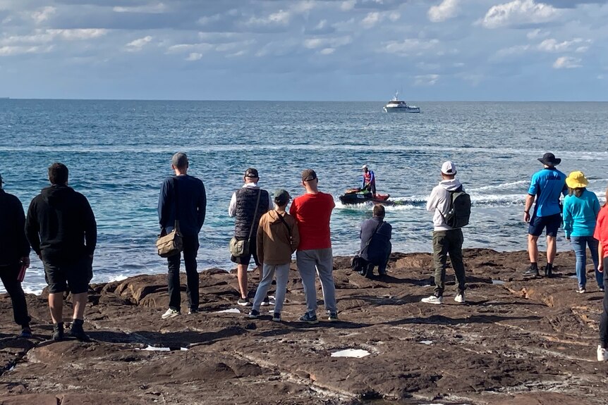 A dozen people standing on rock platform watching a safety briefing in the nearby water.