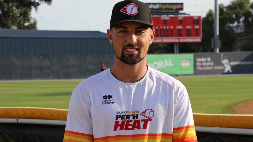 A mid shot of Perth Heat outfielder Jordan Qsar standing in front of a baseball field wearing a heat shirt and cap.