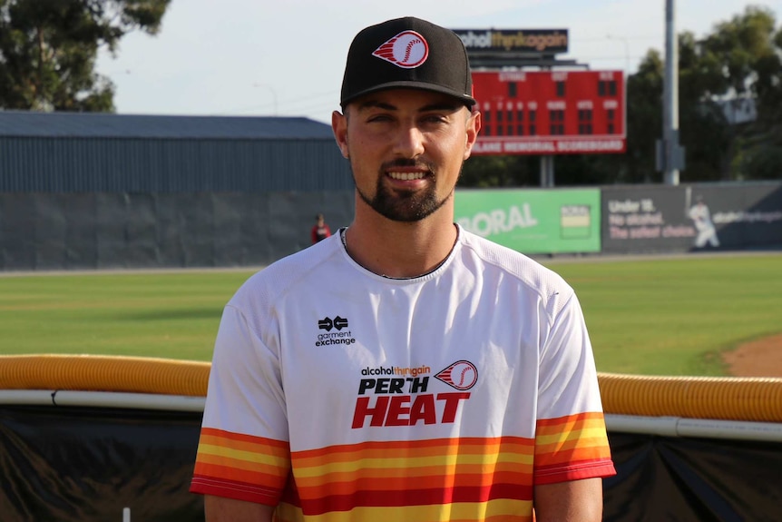A mid shot of Perth Heat outfielder Jordan Qsar standing in front of a baseball field wearing a heat shirt and cap.