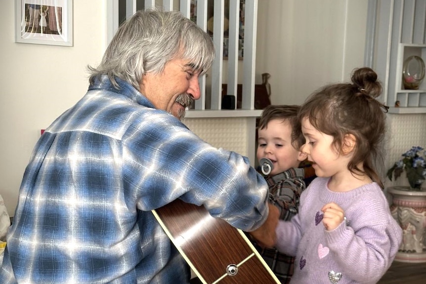 A man wearing a blue, black and white shirt playing the guitar to two young children
