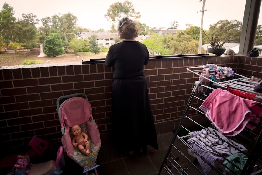 A woman in black clothing on her balcony looking at a park, next to a pink stroller with a toy baby in it.