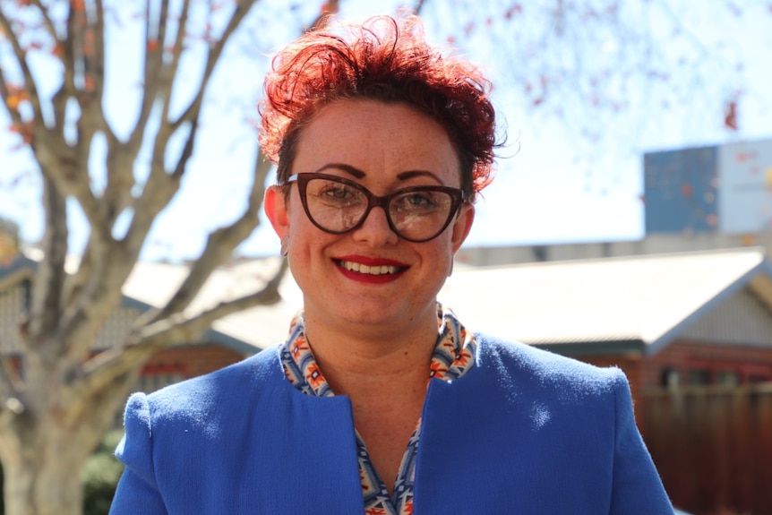 A woman with short red hair wearing glasses and a vivid lilac blazer stands with a tree and house in the background.
