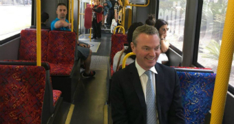 Christopher Pyne sits on a tram with an overly enthusiastic grin. He is on a two-seater chair, but is not near the window.